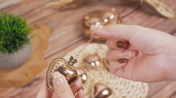 Rose gold tea infuser shaped like a heart, the chain held out so you can see it is long enough to hang off a mug.